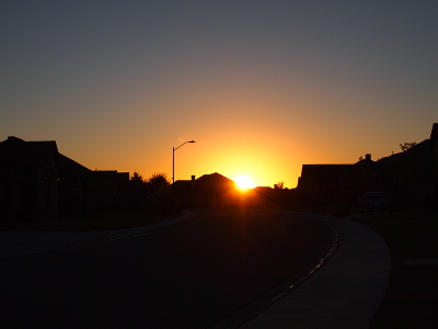 [The sun has just cleared the house rooftops. The rays of the yellow orb brighten the nearby dark home outlines. A half-circle of graduated color from white-yellow to yellow lights the otherwise dark blue sky.]